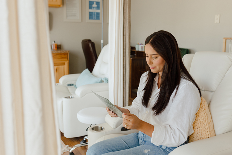 reading-menu-sheet A woman with long dark hair sits in a white armchair, holding a booklet. She is dressed in a white blouse and jeans, and the room has a calming, neutral decor with soft lighting.