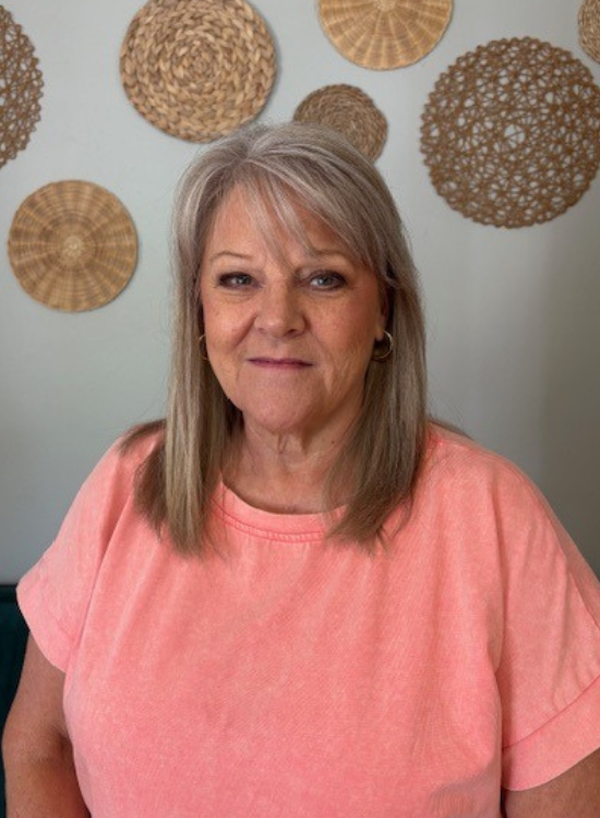 A woman with straight gray hair wearing a peach-colored shirt smiles softly at the camera, standing in front of a wall decorated with round, woven rattan wall hangings.