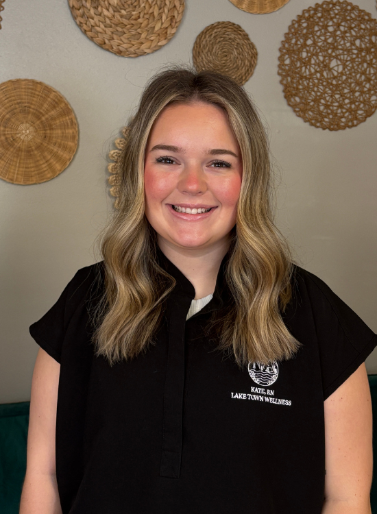 A smiling woman with wavy blonde hair wears a black shirt that reads Lake Town Wellness. She stands in front of a beige wall decorated with round woven baskets.