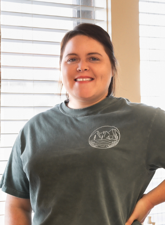A woman with dark hair tied back is smiling and standing indoors in front of window blinds, wearing a green T-shirt with a circular mountain and lake graphic on the chest.