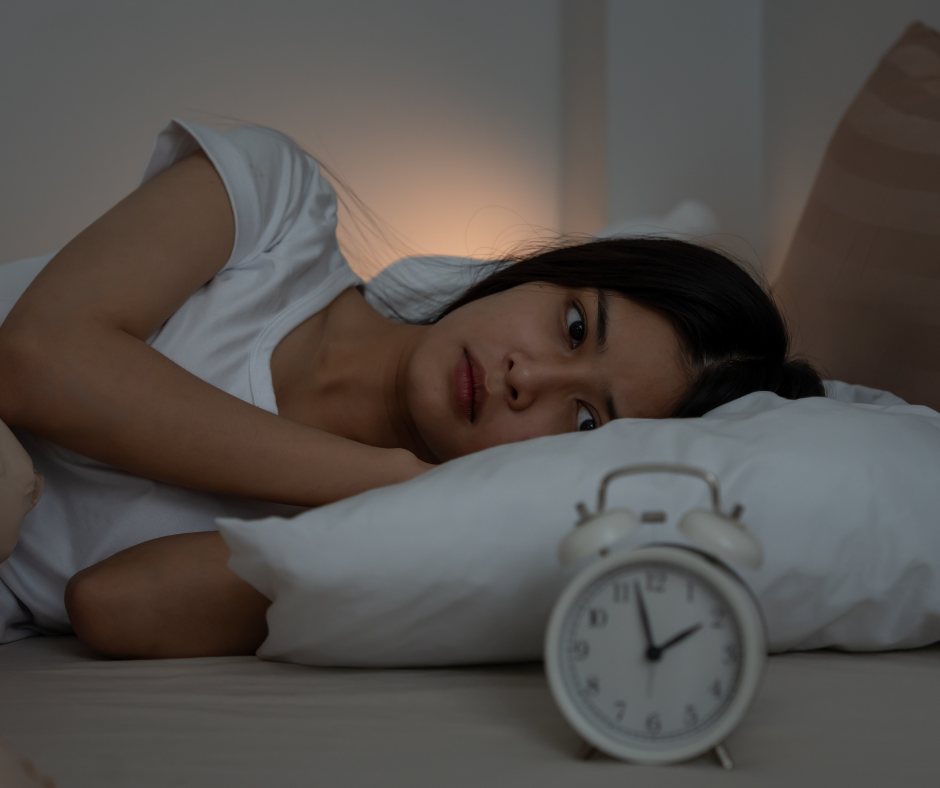 A woman lies in bed at night, looking worried with her head on a pillow. An alarm clock showing 2:10 is on the bedside table in the foreground. The room is dimly lit.