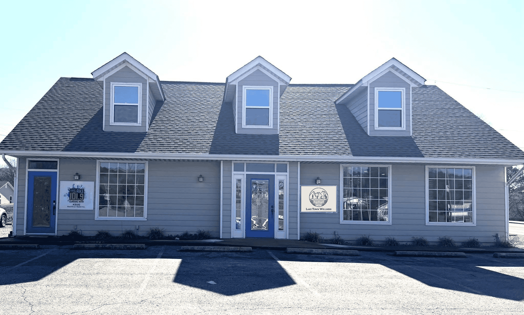 A light-gray, single-story building with three gabled dormers, large front windows, and two business signs near the entrance, situated in a parking lot under a clear sky.