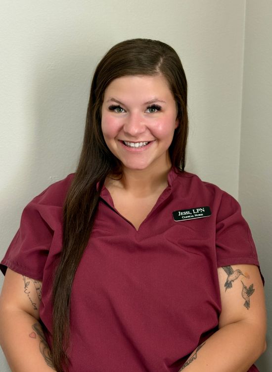 A woman with long brown hair wearing a maroon scrub top and a name badge smiles at the camera. She has visible tattoos on her arms and is seated against a light-colored wall.